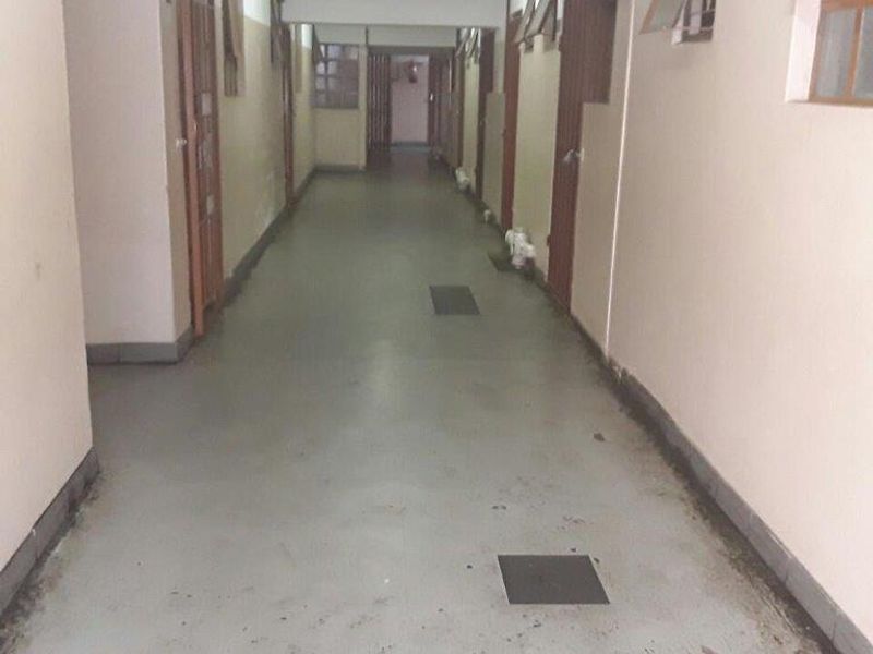 A long apartment building hallway featuring multiple security gates, casement windows, and a fire extinguisher, showing signs of wear and maintenance requirements.