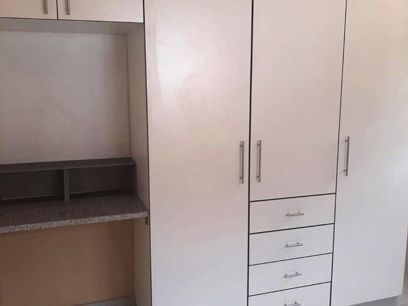 A bedroom featuring white built-in cupboards with integrated drawers and a built-in desk area, finished with neutral floor tiles.