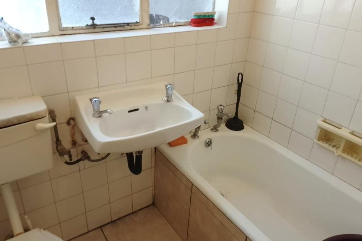 A dated bathroom featuring a wall-mounted basin, bathtub, and toilet, with tiled finishes and visible plumbing requiring maintenance or renovation.