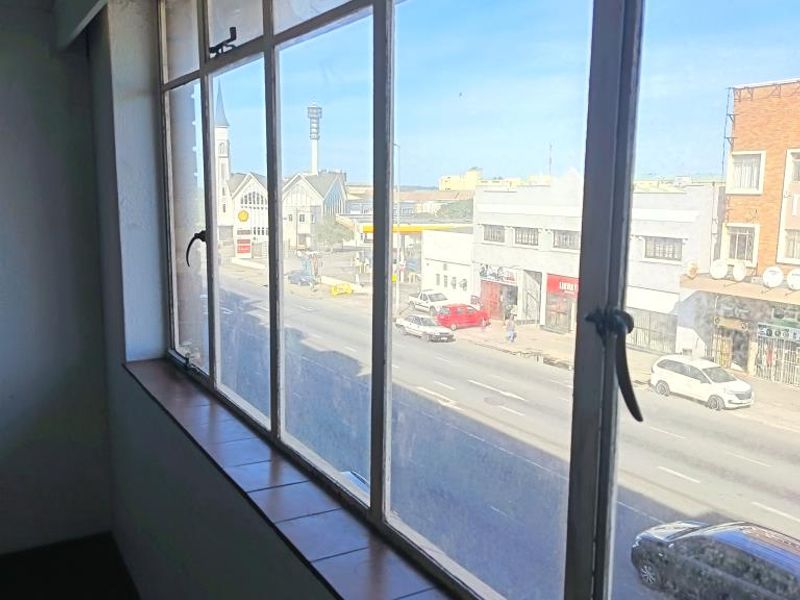 An interior view looking out through large metal-framed windows onto a street scene with surrounding urban buildings.