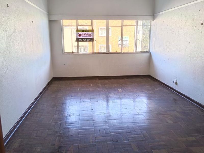 An empty living room featuring original parquet flooring, a large window, and a ceiling light fixture, requiring cosmetic updates to the walls.