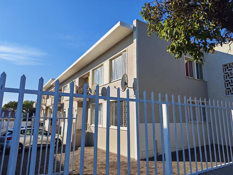 Exterior view of a multi-story apartment building featuring a paved driveway, security fencing, and private balconies.