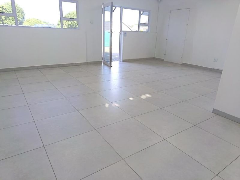 A bright, open-plan commercial office space featuring neutral tiled flooring, large windows, and direct access to an outdoor area.