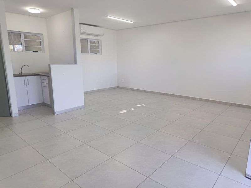 A bright, open-plan commercial space featuring neutral tiled flooring, a compact kitchenette with sink, and a wall-mounted air conditioning unit.