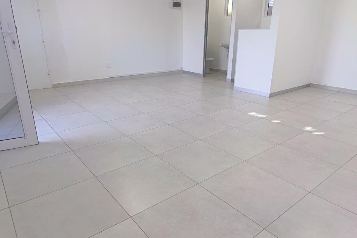 A bright, vacant open-plan commercial space featuring neutral tiled flooring, clean white walls, and direct access to a private restroom facility.