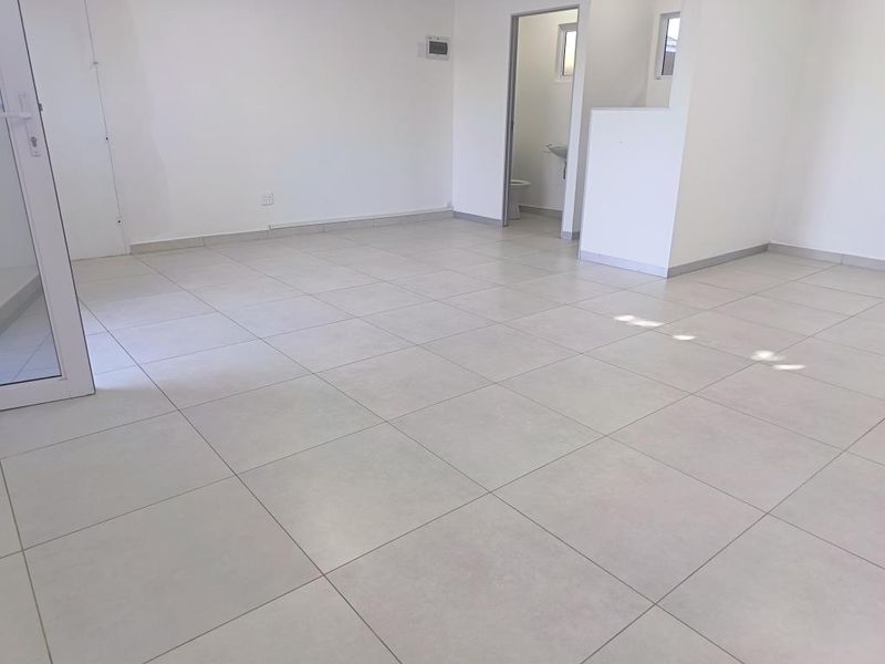 A bright, vacant open-plan commercial space featuring neutral tiled flooring, clean white walls, and direct access to a private restroom facility.