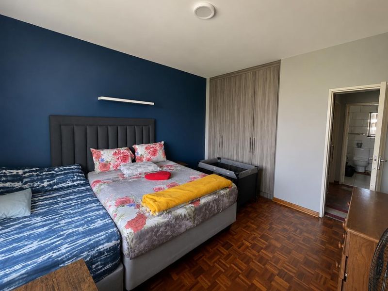 Spacious bedroom featuring warm parquet flooring, a striking navy accent wall, and ample built-in cupboard storage, with direct access to an en-suite bathroom.