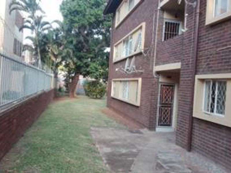 Rear view of a multi-story brick apartment building featuring security gates, window bars, and a shared garden area with perimeter fencing.