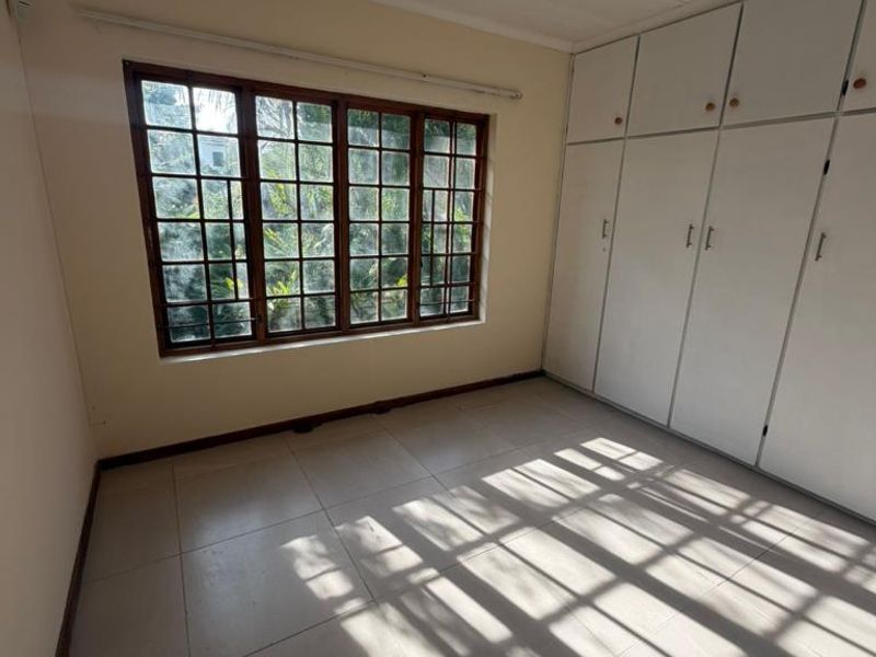 A bright, empty bedroom featuring tiled flooring, built-in cupboards, and a large wooden-framed window allowing for natural light.