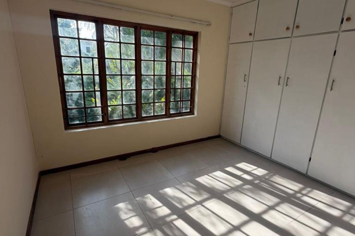 A bright, empty bedroom featuring tiled flooring, built-in cupboards, and a large wooden-framed window allowing for natural light.