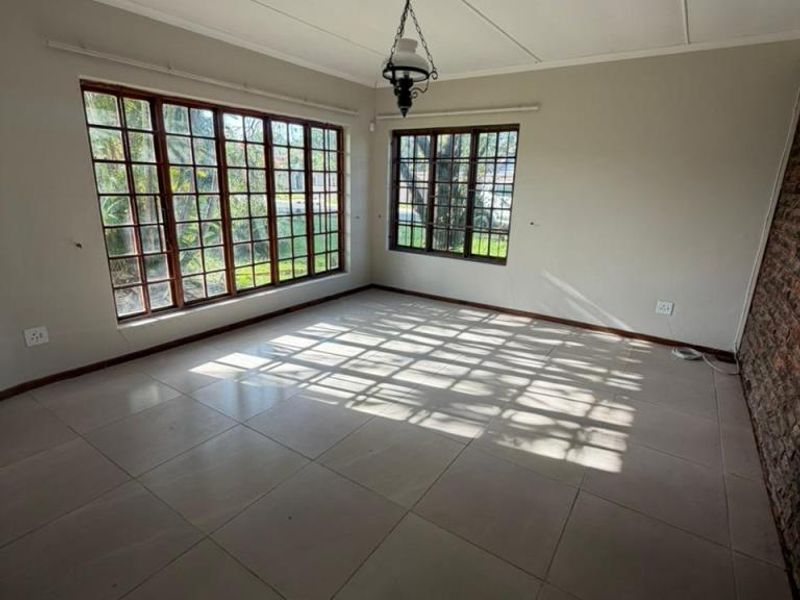 A bright, empty living room featuring large multi-pane windows, tiled flooring, and an exposed brick feature wall, offering a versatile blank canvas.