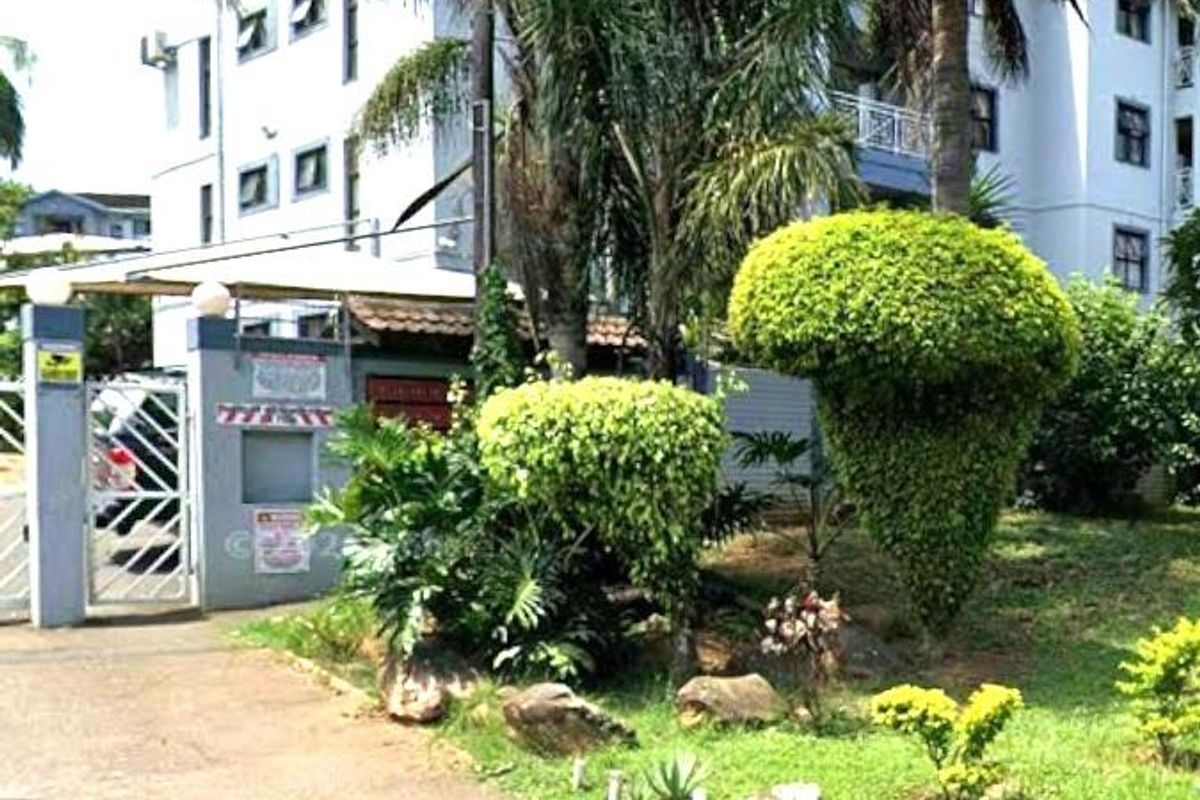Front exterior view of the apartment complex featuring a gated entrance, paved driveway, and established garden landscaping.