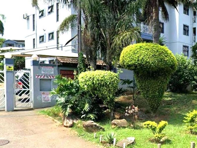 Front exterior view of the apartment complex featuring a gated entrance, paved driveway, and established garden landscaping.