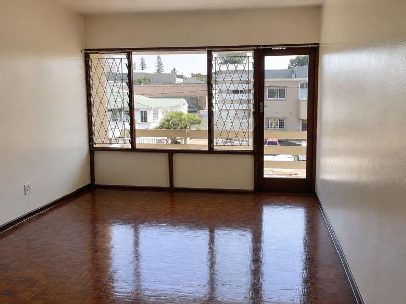 An empty living room featuring polished parquet flooring, large windows with security bars, and direct access to a balcony.