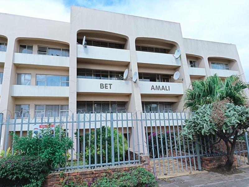 Front elevation of the Bet Amali apartment building, featuring multi-level balconies, security fencing, and established garden landscaping.