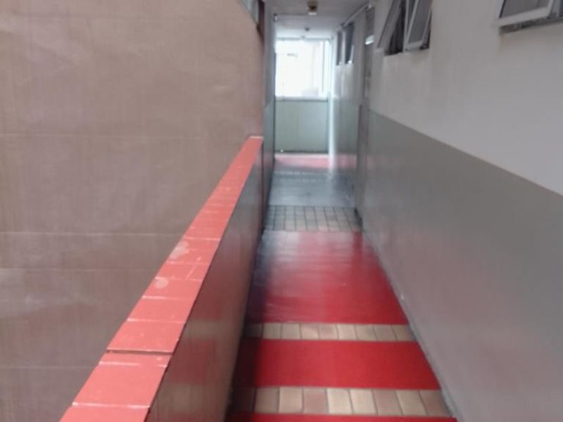 An open-air apartment building corridor featuring tiled flooring, a red balustrade, and side-facing casement windows.