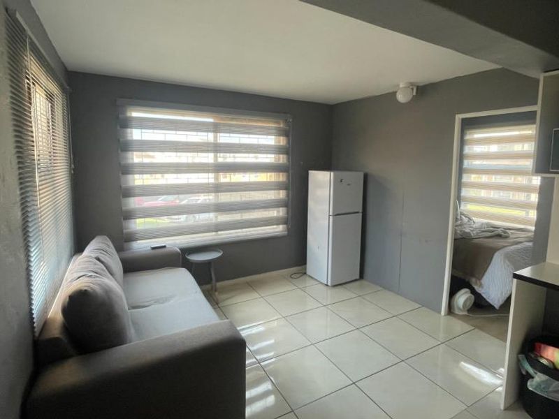 Open plan living area featuring tiled flooring, neutral grey walls, and large windows with fitted roller blinds and security bars.