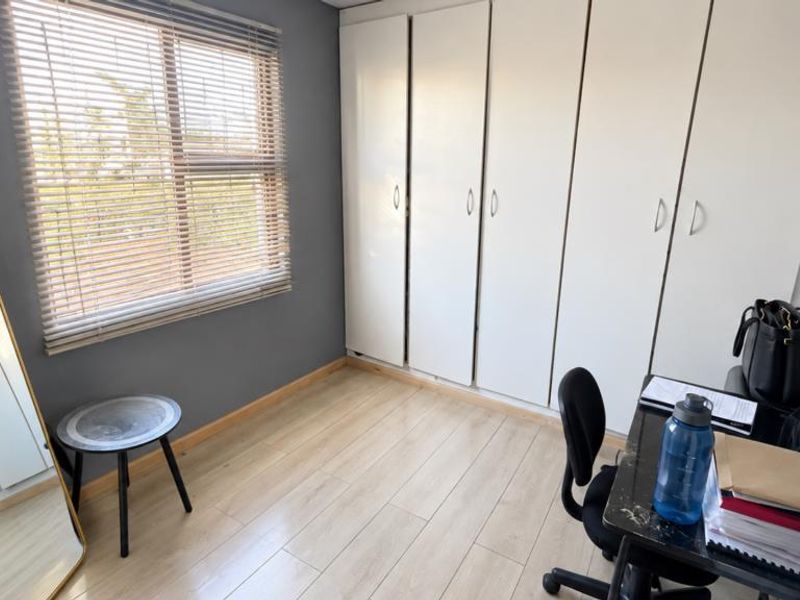 A bedroom featuring light-toned laminate flooring, built-in white cupboards, and a window fitted with venetian blinds.