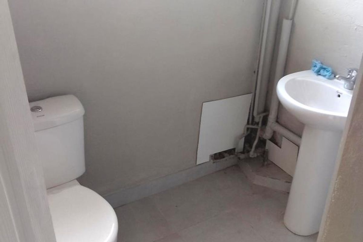 A compact bathroom featuring a pedestal basin, standard toilet, and exposed plumbing, with security bars on the window.