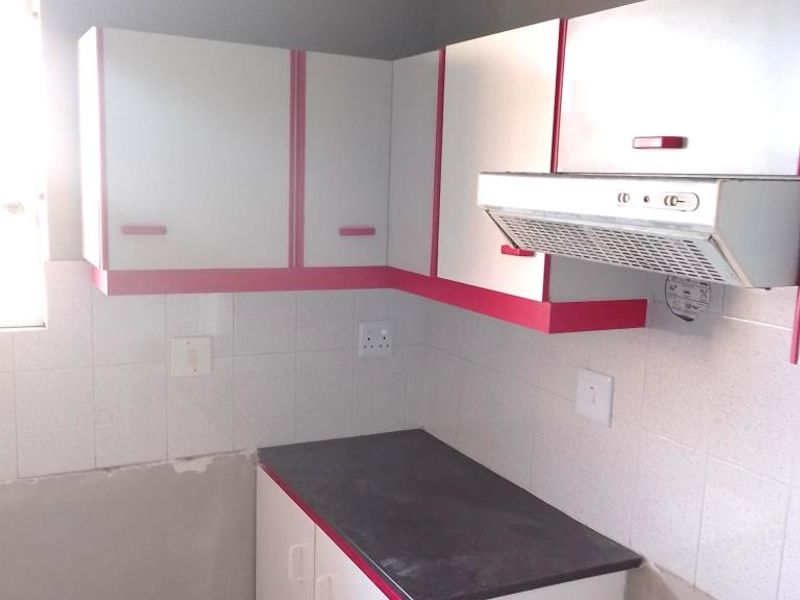 A compact kitchen featuring white and pink cabinetry, a dark countertop, and an extractor fan, currently requiring finishing work on the lower wall sections.