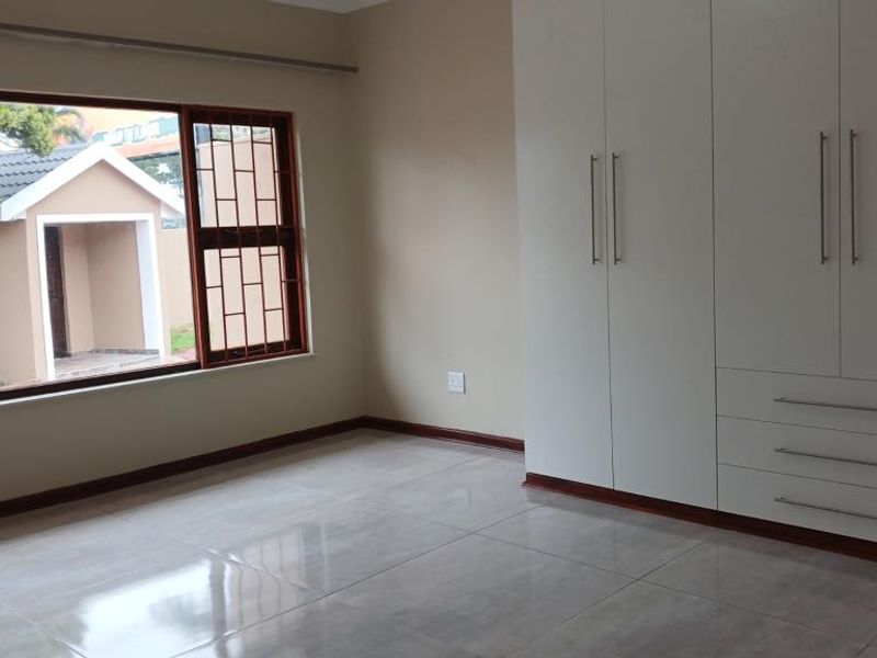 A bright, empty bedroom featuring modern white built-in cupboards, large windows with security bars, and neutral tiled flooring.