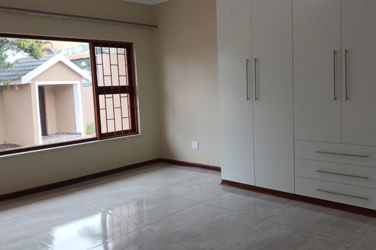 A bright, empty bedroom featuring modern white built-in cupboards, large windows with security bars, and neutral tiled flooring.