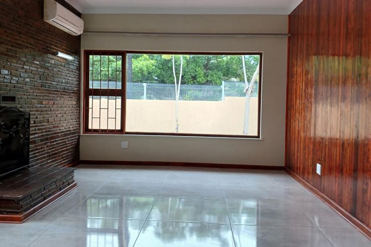 Spacious living room featuring a brick fireplace, wood-paneled accent wall, large window with security bars, and polished tiled flooring.