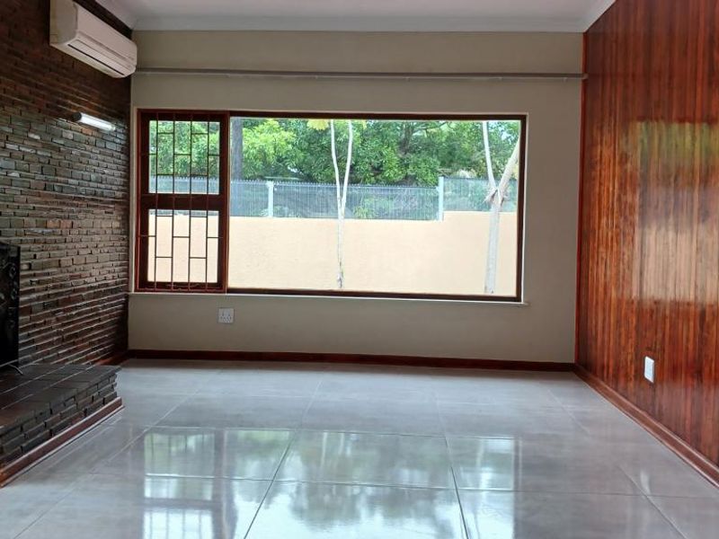 Spacious living room featuring a brick fireplace, wood-paneled accent wall, large window with security bars, and polished tiled flooring.