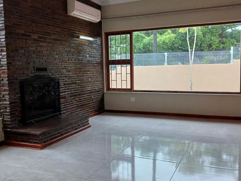 Spacious living room featuring a prominent brick fireplace, large picture window, tiled flooring, and a wall-mounted air conditioning unit.