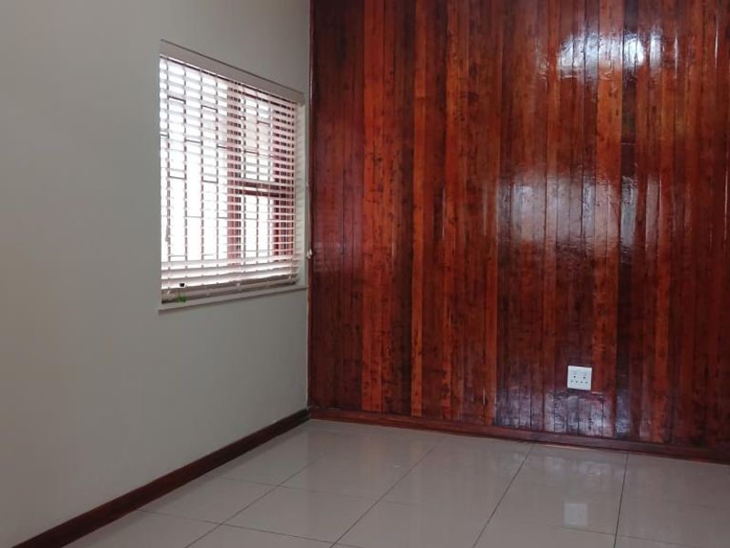 A clean, empty bedroom featuring polished wooden wall paneling, neutral tiled flooring, and a window fitted with venetian blinds.