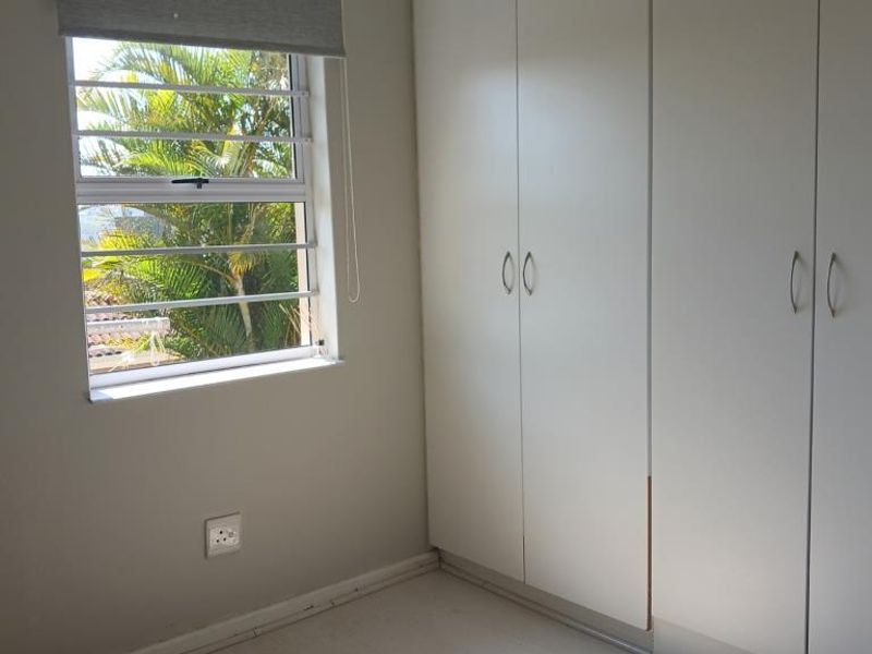 A bright, empty bedroom featuring built-in cupboards, a window with security bars, and a roller blind.
