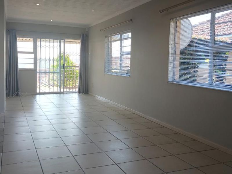 A spacious, empty living area featuring neutral tiled flooring, recessed lighting, and large windows with security gates and blinds, offering a bright, blank canvas.