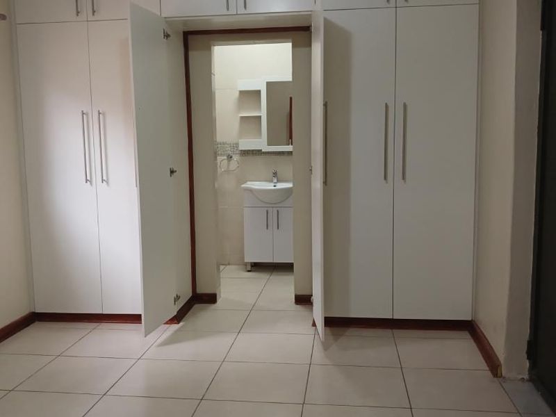 A clean, empty bedroom featuring extensive built-in cupboards, tiled flooring, and direct access to an en-suite bathroom with a vanity basin.
