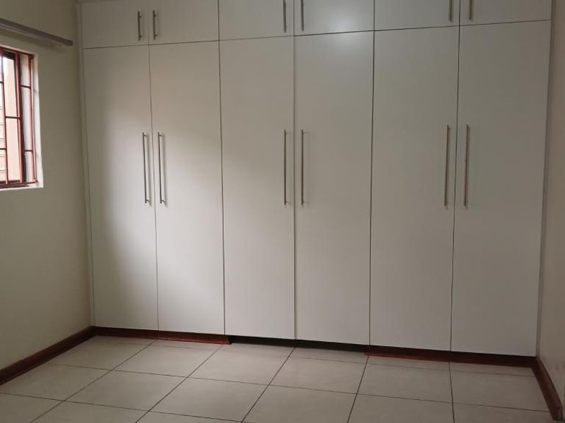 A clean, empty bedroom featuring extensive white built-in cupboards, tiled flooring, and a window fitted with security burglar bars.
