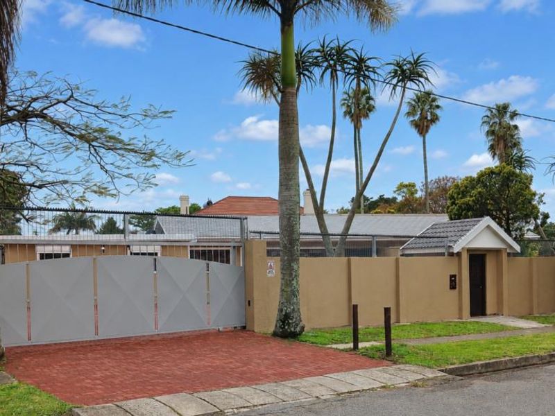 Front elevation of the property featuring a secure perimeter wall, paved driveway, and gated entrance with established landscaping.