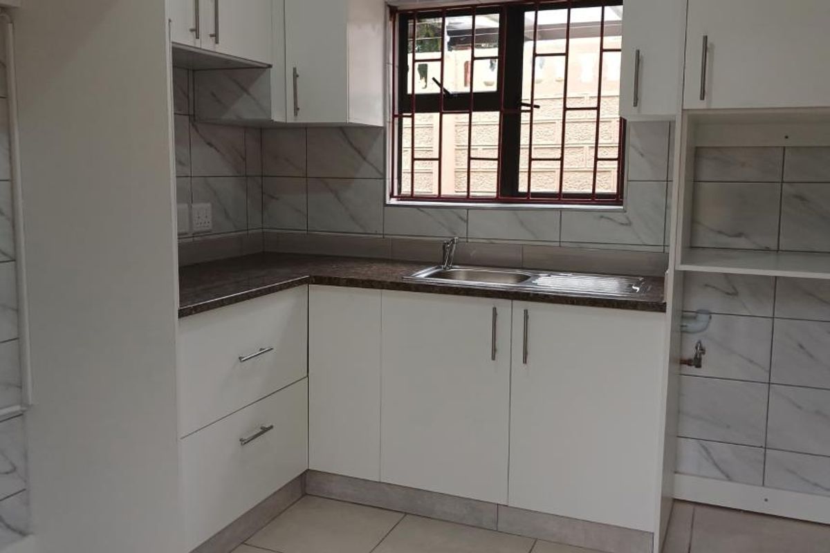 A clean, modern kitchen featuring white built-in cabinetry, marble-look tiled backsplash, laminate countertops, and a stainless steel sink with window security bars.