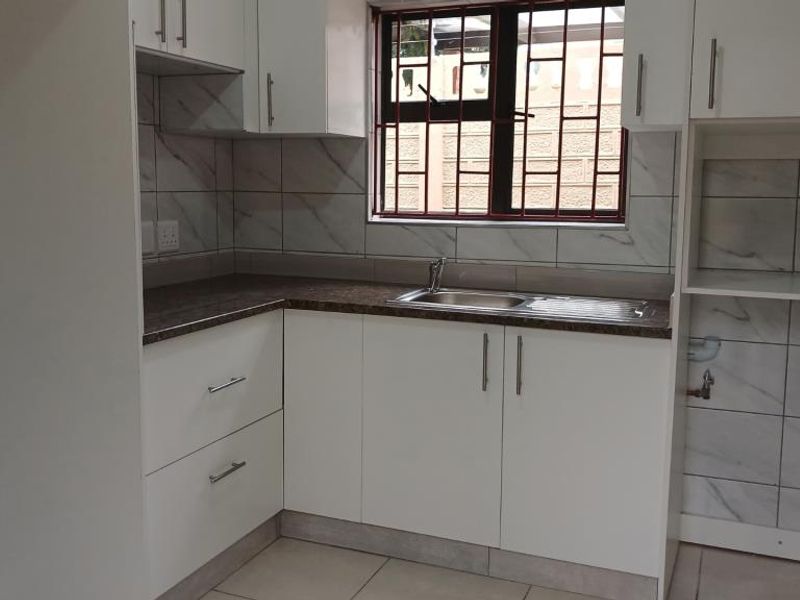 A clean, modern kitchen featuring white built-in cabinetry, marble-look tiled backsplash, laminate countertops, and a stainless steel sink with window security bars.