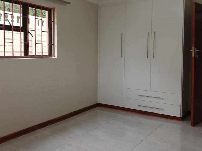 A clean, empty bedroom featuring modern white built-in cupboards, neutral tiled flooring, and a window fitted with security bars.