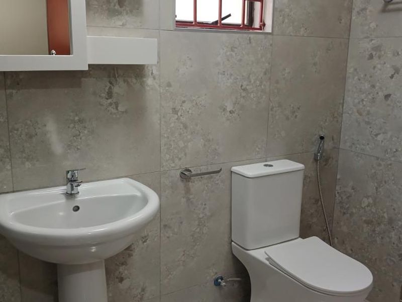 A clean, modern bathroom featuring neutral stone-look wall tiles, a white pedestal basin, and a standard toilet with a bidet spray attachment.