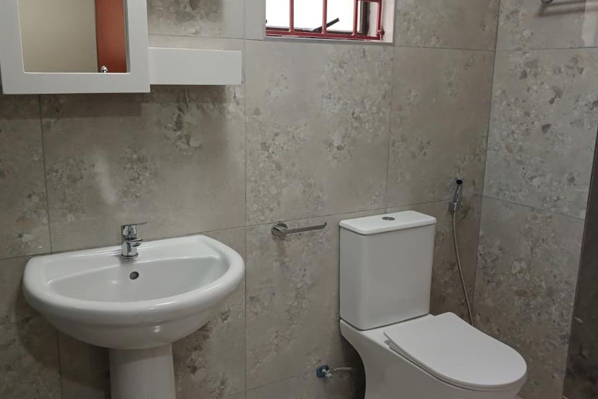 A clean, modern bathroom featuring neutral stone-look wall tiles, a white pedestal basin, and a standard toilet with a bidet spray attachment.