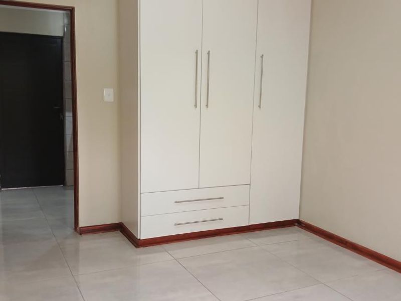 A clean, empty bedroom featuring neutral wall tones, large format floor tiles, and a modern white built-in wardrobe with integrated drawers.