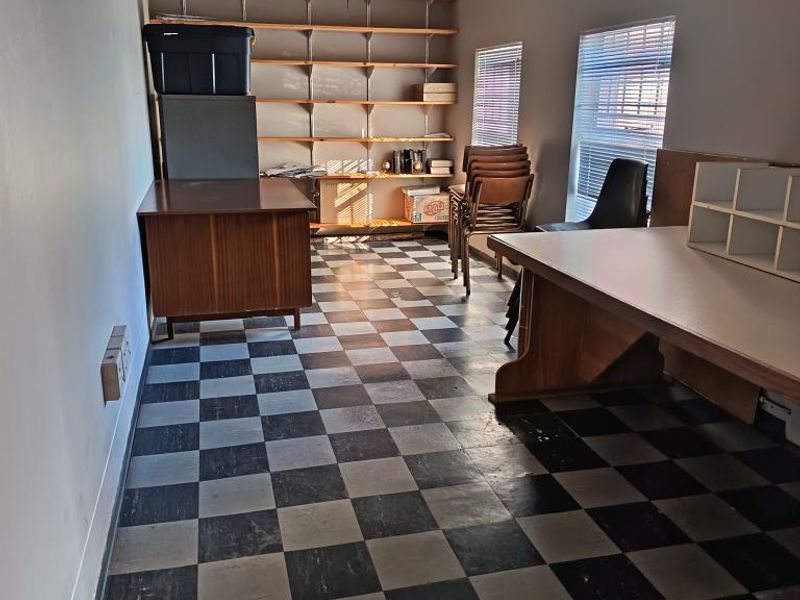 A functional office space featuring checkerboard vinyl flooring, wall-mounted shelving, and natural light through venetian blinds.