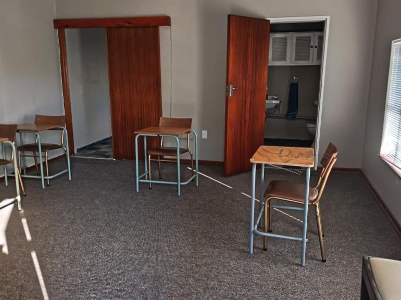 A functional office space featuring carpeted flooring, dual access doors, and an adjoining small bathroom area with built-in cabinetry.