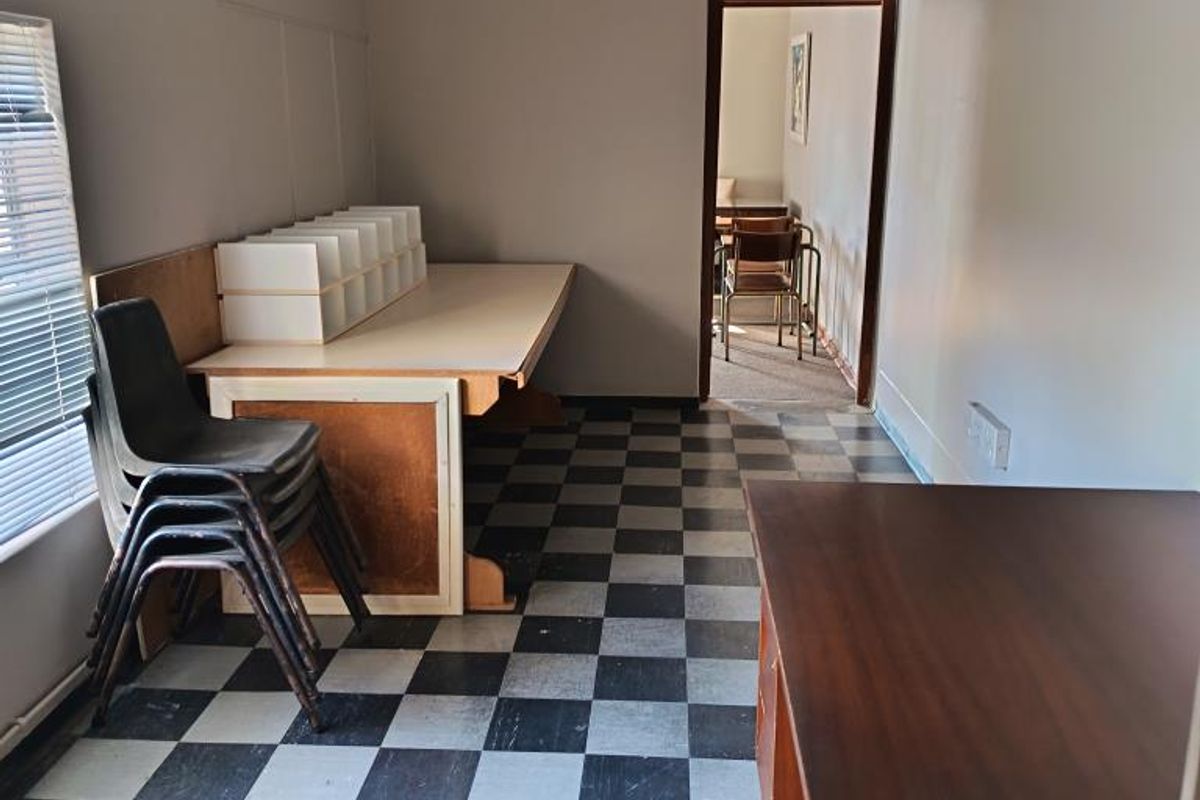 A functional office space featuring checkerboard vinyl flooring, venetian blinds, and direct access to an adjoining room.