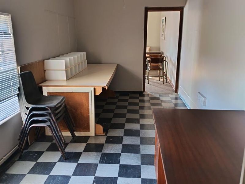 A functional office space featuring checkerboard vinyl flooring, venetian blinds, and direct access to an adjoining room.