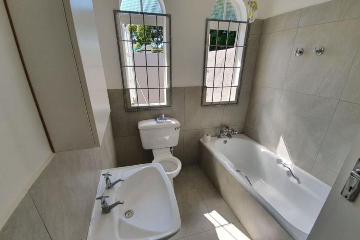 A functional bathroom featuring a bathtub, pedestal basin, toilet, and neutral wall and floor tiling with security bars on the windows.
