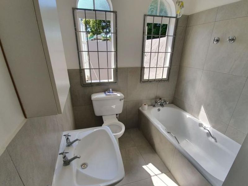A functional bathroom featuring a bathtub, pedestal basin, toilet, and neutral wall and floor tiling with security bars on the windows.