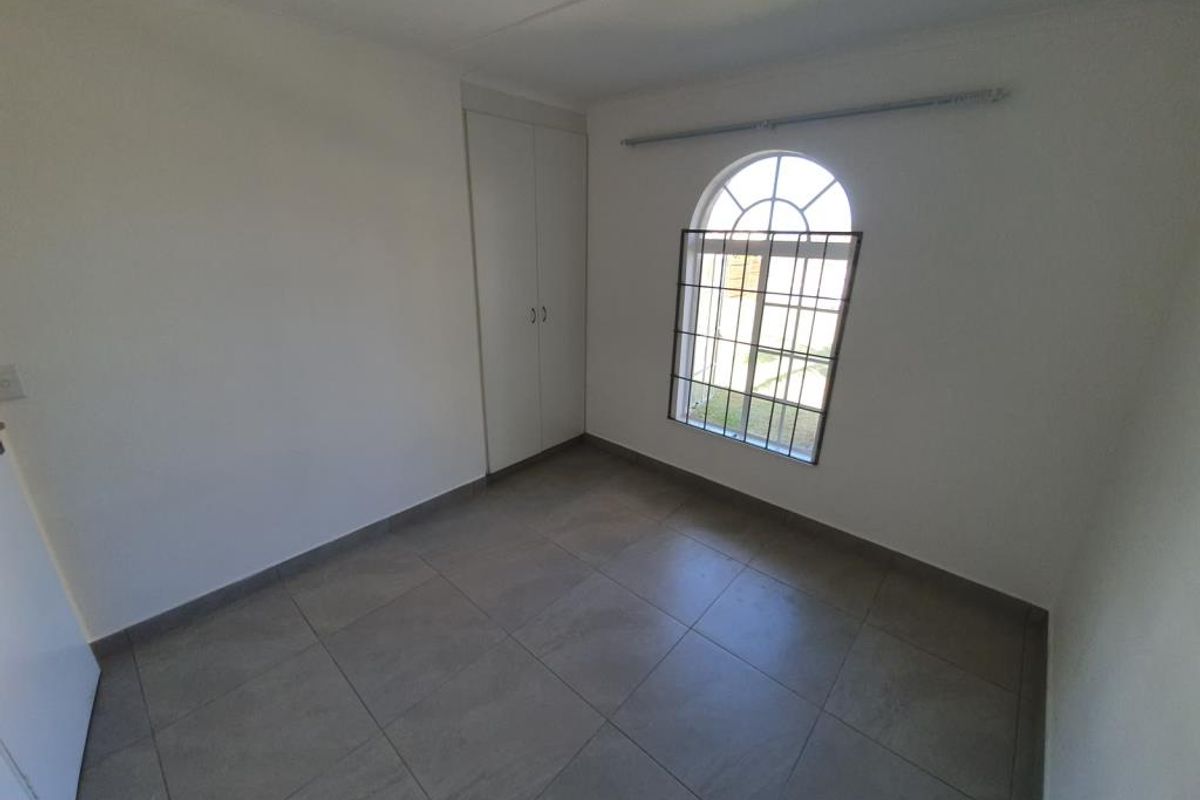 A bright, empty bedroom featuring neutral tiled flooring, built-in cupboards, and an arched window with security bars.