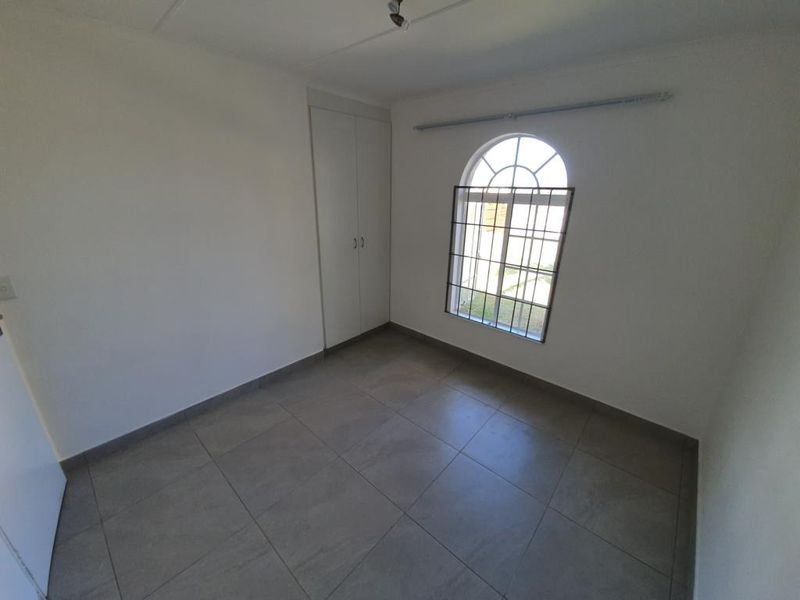 A bright, empty bedroom featuring neutral tiled flooring, built-in cupboards, and an arched window with security bars.
