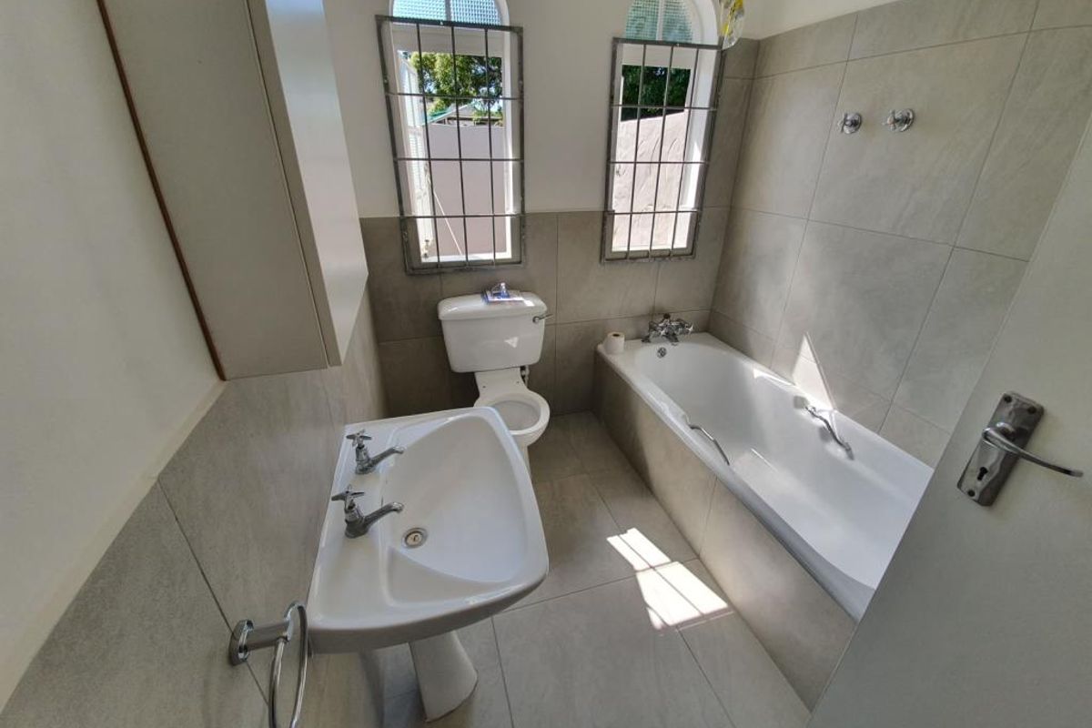 A functional bathroom featuring a pedestal basin, toilet, and bathtub with neutral wall and floor tiling and security-fitted windows.